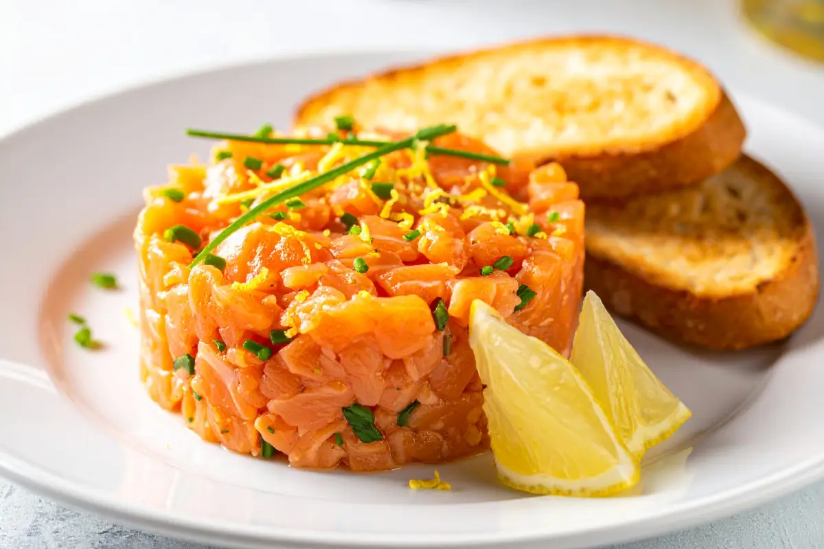 Salmon tartare served in a round mold on a white plate, garnished with fresh chives and lemon zest, accompanied by toasted bread slices, French brasserie style.